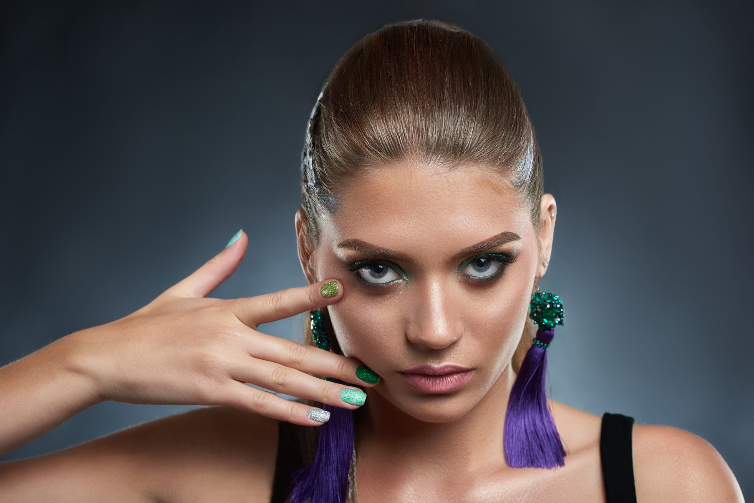 Studio portrait of attractive and seductive woman with shiny manicure and makeup in green colors. Beautiful brunette with long violet earring looking at camera, touching face. Beauty concept.